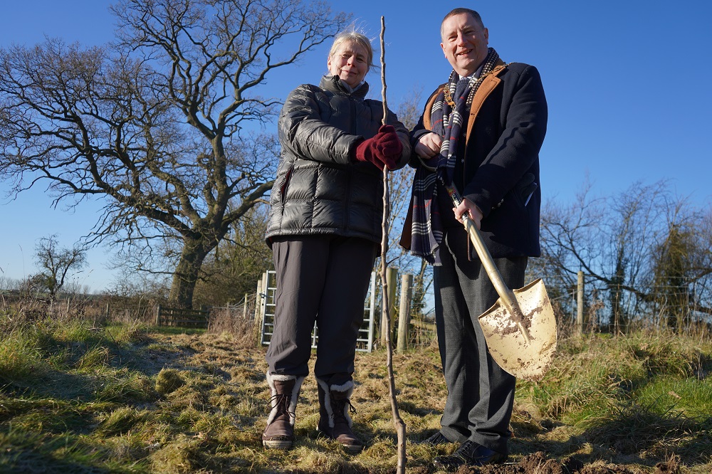 Two people stood in front of a young tree with a spade.